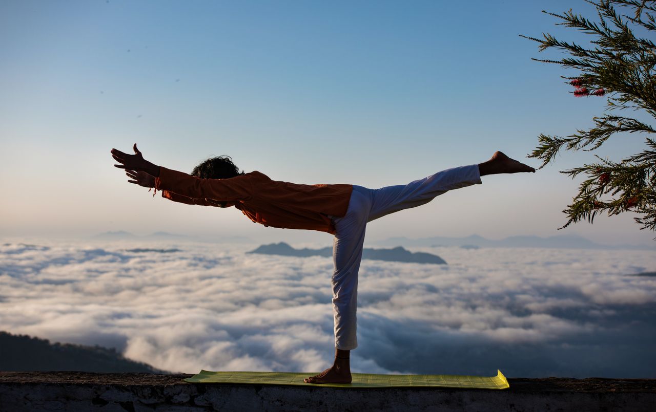 hero-img-01 Person practicing yoga in warrior pose above a scenic cloudscape during sunrise.